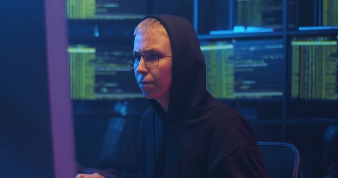 Caucasian cyberpolice woman in glasses and black hood sitting at computer screen as working at night and turning to look at data on monitors behind. Female cyber criminal stealing information online.
