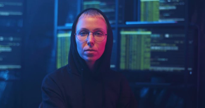 Caucasin female hacker in dark hood and glasses turning to camera on spinning chair and looking straight at night in room with monitors and data. Portrait of woman sittng in cyber police offce.