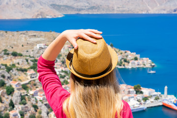 Girl with wavy hair in straw hat and pink long sleeves shirt is standing on top of the mountain and watching down on tiny colorful houses and Mediterranean sea on greek island Symi