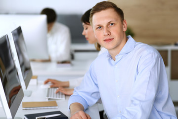 Fototapeta premium Cheerful smiling businessman headshot at work in modern office. Unknown casual dressed entrepreneur using pc computer while sitting with diverse colleagues at the background. Multi ethnic working