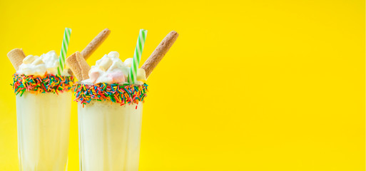 Milkshake with ice cream and whipped cream, marshmallows, waffles, served in a glass cup decorated with pastry dressing. Summer cocktail. Bright yellow background. Banner. Copy space.