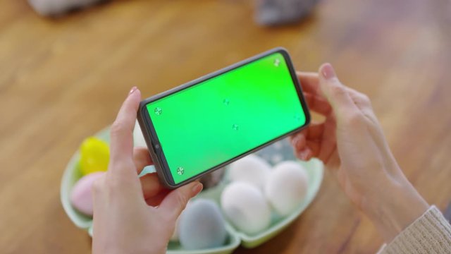 Handheld close up shot of female hands holding mobile phone with chroma key screen over tray of dyed Easter eggs