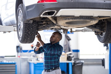 African male car mechanic standing under the car on the lift in the service center, inspect the...