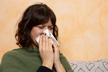 Nice brown haired female sitting on couch and blowing her nose to paper tissue. Tired woman...
