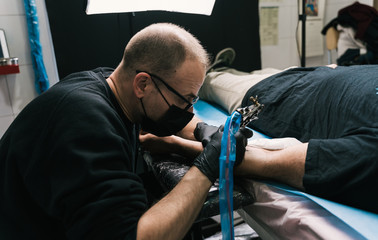 A closeup shot of a tattoo artist with face mask creating a tattoo on a man's arm
