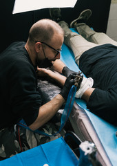 A high angle shot of a tattoo artist with eyeglasses creating a tattoo on a man's arm