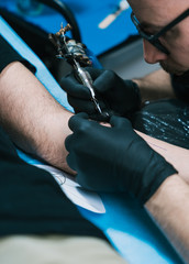 A closeup shot of a tattoo artist with eyeglasses creating a tattoo on a man's arm
