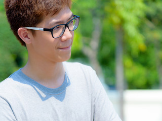 Asian handsome teenage boy wearing glasses and thoughtful gestures, Portrait cheerful hipster young man pursed lips and looking with a suspicious face at park on green nature background in Thailand
