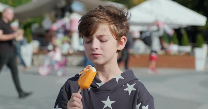 Portrait happy boy eats an orange popsicle on stick with great pleasure against central city square and people walking sunny day autumn. Delicious Ice Cream. Emotions. Lifestyle