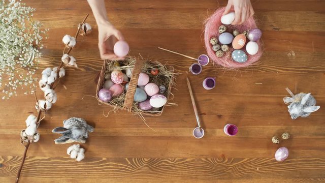 Top view of female hands taking colorful Easter eggs out of handmade pink nest and putting them into basket with straw