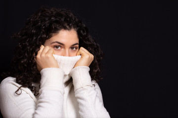 Pretty young brunette with curly long hair pulling up a white woolen turtleneck sweater over her mouth for warmth. Winter cold season concept