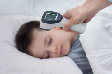 Na pierwszym planie dłoń trzyma termometr elektroniczny gotowy do pomiaru.  W tle twarz chłopca śpiącego w łóżku na białej pościeli. 