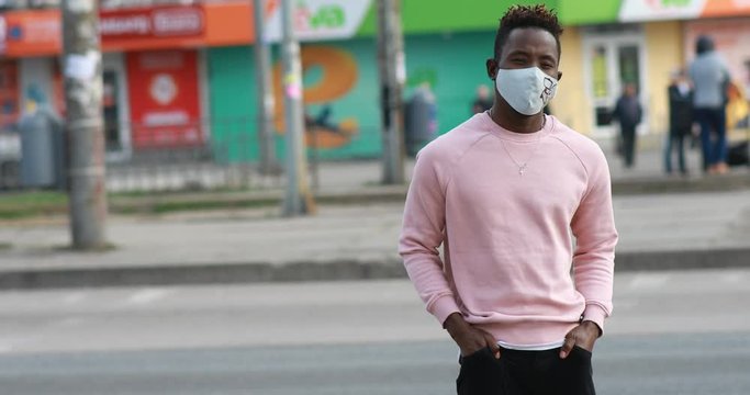 A young African man stands with a medical protective mask on his face at city street. Concept of preventive measures and protection for coronavirus pandemic. 
