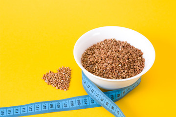 raw buckwheat in a small white bowl, blue measuring tape and buckwheat heart on a yellow background copy space, proper nutrition concept