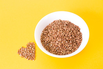 raw buckwheat in a small white plate and a heart of buckwheat on a yellow background copy space