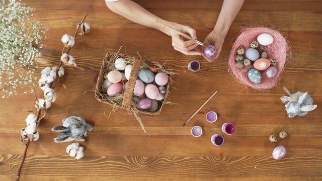 Top view of female hands painting pattern on dyed Easter egg. Basket with colorful eggs, bunny figurines and flowers standing on wooden table