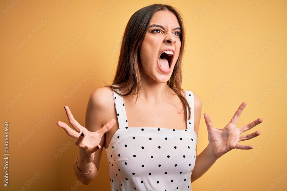 Wall mural young beautiful brunette woman wearing casual dress standing over yellow background crazy and mad sh