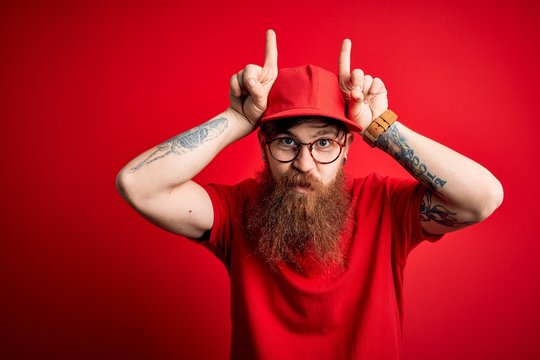 Young handsome delivery man wearing glasses and red cap over isolated background doing funny gesture with finger over head as bull horns
