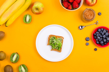 Toast with peanut butter and kiwi on a white plate among fruits and berries on a bright yellow background. Top view, flat lay.