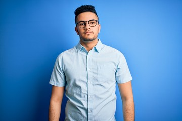 Fototapeta premium Young handsome man wearing casual summer shirt and glasses over isolated blue background with serious expression on face. Simple and natural looking at the camera.