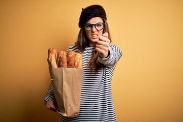 Young beautiful brunette french woman wearing beret holding paper bag with baguettes annoyed and frustrated shouting with anger, crazy and yelling with raised hand, anger concept