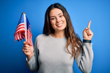 Young beautiful brunette patriotic woman holding american flag celebrating 4th of july surprised...