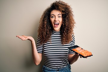 Young beautiful woman with curly hair and piercing holding tray with fresh sushi very happy and excited, winner expression celebrating victory screaming with big smile and raised hands