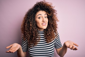 Young beautiful woman with curly hair and piercing wearing casual striped t-shirt clueless and...