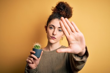 Young beautiful woman with curly hair and piercing holding pot with cactus plant with open hand...