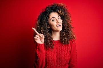 Young beautiful woman with curly hair and piercing wearing casual red sweater pointing finger up with successful idea. Exited and happy. Number one.