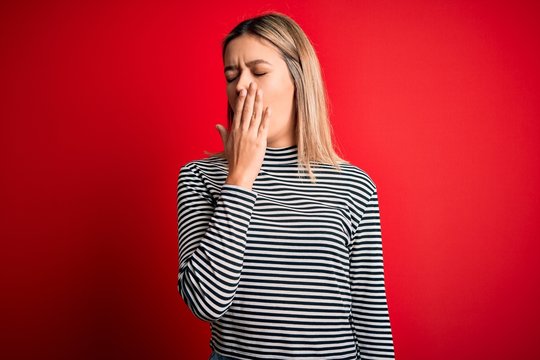 Young beautiful blonde woman wearing casual striped sweater over red isolated background bored yawning tired covering mouth with hand. Restless and sleepiness.