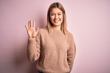 Young beautiful blonde woman wearing winter wool sweater over pink isolated background showing and pointing up with fingers number five while smiling confident and happy.