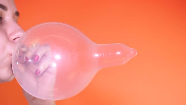 A woman inflates a condom on an orange background, close-up.
