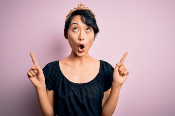 Young beautiful chinese woman wearing golden crown of king over isolated pink background amazed and surprised looking up and pointing with fingers and raised arms.