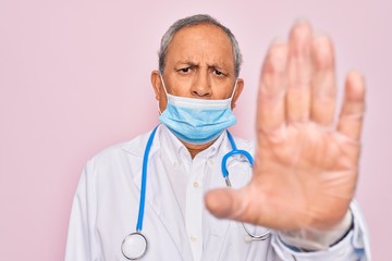 Senior hoary doctor man wearing medical mask and stethoscope over pink background with open hand doing stop sign with serious and confident expression, defense gesture