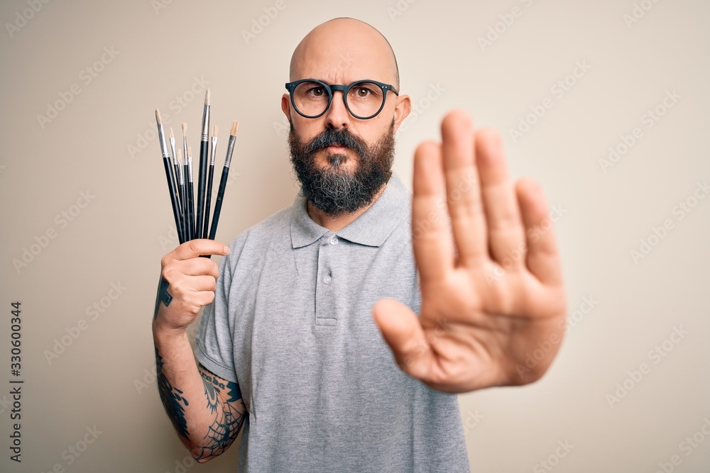 Wall mural Handsome bald artist man with beard and tattoo painting using painter brushes with open hand doing stop sign with serious and confident expression, defense gesture