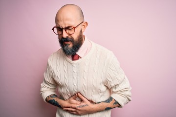 Handsome bald man with beard and tattoo wearing glasses and sweater over pink background with hand on stomach because nausea, painful disease feeling unwell. Ache concept.