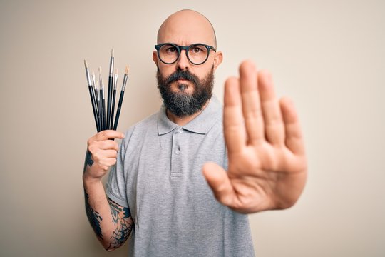 Handsome bald artist man with beard and tattoo painting using painter brushes with open hand doing stop sign with serious and confident expression, defense gesture