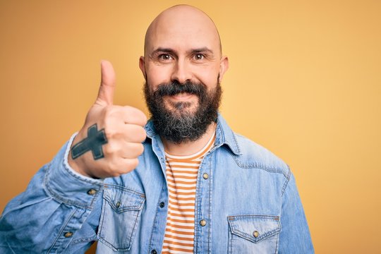 Handsome bald man with beard wearing casual denim jacket and striped t-shirt doing happy thumbs up gesture with hand. Approving expression looking at the camera showing success.