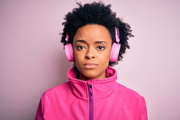 Young African American afro woman with curly hair listening to music using pink headphones with serious expression on face. Simple and natural looking at the camera.