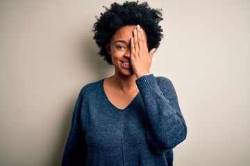 Fototapeta na wymiar Young beautiful African American afro woman with curly hair wearing casual sweater covering one eye with hand, confident smile on face and surprise emotion.