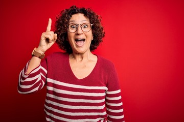 Middle age beautiful curly hair woman wearing casual striped sweater over red background pointing finger up with successful idea. Exited and happy. Number one.