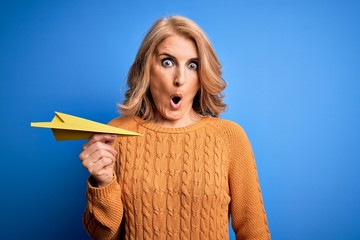 Middle age beautiful blonde woman holding paper airplane over isolated blue background scared in...