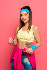 beautiful sportswoman holding dumbbells while looking at camera on pink background
