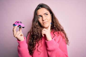 Young beautiful woman with curly hair holding small car over isolated pink background serious face thinking about question, very confused idea