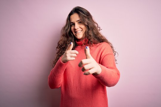 Young beautiful woman with curly hair wearing turtleneck sweater over pink background pointing fingers to camera with happy and funny face. Good energy and vibes.