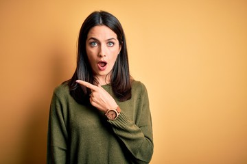 Young brunette woman with blue eyes wearing green casual sweater over yellow background Surprised pointing with finger to the side, open mouth amazed expression.