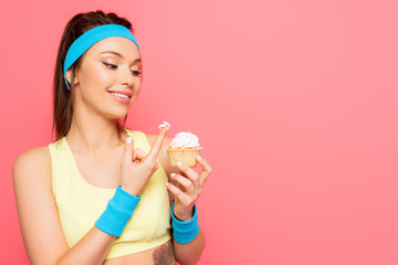cheerful sportswoman with whipped cream on finger holding delicious cupcake isolated on pink