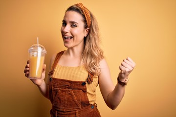 Young beautiful blonde woman drinking healthy glass of orange juice over yellow background screaming proud and celebrating victory and success very excited, cheering emotion