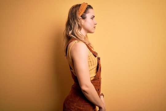Young beautiful blonde woman wearing overalls and diadem standing over yellow background looking to side, relax profile pose with natural face and confident smile.